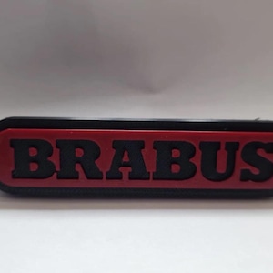 Puede incluir: Un emblema rectangular con la palabra "BRABUS" en letras negras en negrita. Las letras están sobre un fondo rojo, enmarcado por un borde negro. El emblema es probablemente para uso automotriz.