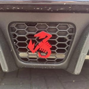 Puede incluir: Primer plano de un parachoques de coche negro con una rejilla de panal. Un emblema de escorpión rojo brillante está centrado en la rejilla. El emblema es un escorpión estilizado con la cola curvada hacia arriba. El parachoques está ligeramente inclinado.