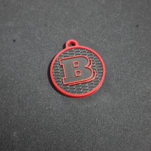 Op de afbeelding: Een ronde, rode en zwarte sleutelhanger met de letter "B" in het midden. De letter is zwart, en de achtergrond heeft een honingraatpatroon. De sleutelhanger heeft een kleine lus aan de bovenkant.