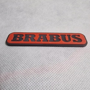 Può includere: Un emblema rettangolare con la scritta "BRABUS" in lettere nere in rilievo. Le lettere sono su uno sfondo rosso, con un bordo nero. L'emblema è su una superficie bianca strutturata.