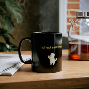 Könnte beinhalten: Schwarze Keramik-Tasse mit goldener Aufschrift und einer Cartoon-Katzen-Illustration. Die Tasse hat einen gebogenen Henkel und steht auf einer Holzoberfläche. Auf der Tasse steht der Text "Trinke Tee und sei nicht traurig".