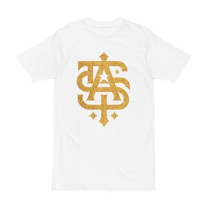 May include: White t-shirt featuring a gold-colored monogram design. The monogram is an ornate arrangement of the letters "T", "A", and "S", with a decorative pattern. The shirt has short sleeves and a crew neck.