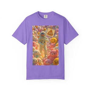 May include: A lilac-colored t-shirt featuring a whimsical graphic. The graphic depicts an astronaut holding a sign with a poop emoji, surrounded by plush toys. The astronaut is wearing a white spacesuit. The plush toys are various colors.