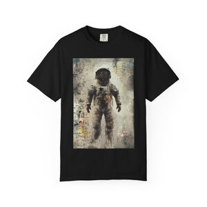 May include: Black t-shirt featuring a full-body illustration of an astronaut in a spacesuit. The artwork is in muted tones with visible brushstrokes and abstract elements. The astronaut is the central focus, set against a textured background.