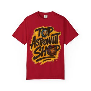 May include: A red t-shirt with the words "TOP ASTRONAUT SHOP" in black and yellow, with illustrations of astronauts in helmets. The design has a retro, graffiti-style aesthetic.