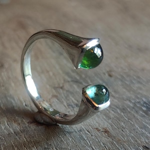 Op de afbeelding: Een open zilveren ring met twee gepolijste, ronde groene edelstenen aan beide uiteinden van de band. De metalen ring heeft een gladde, reflecterende afwerking en de twee stenen hebben licht verschillende tinten groen, een donkere en een lichtere.
