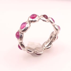 Peut inclure: Une bague en argent ornée d'une série de pierres précieuses ovales de couleur magenta disposées autour du cercle. La bague est marquée "925" à l'intérieur. Les pierres précieuses sont espacées uniformément.