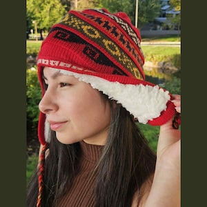 May include: A red and patterned winter hat with a white, fluffy lining. The hat features a geometric design in black, yellow, and green. The hat has ear flaps and braided tassels with red and yellow threads.