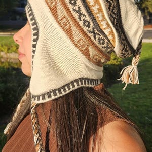 May include: A knitted hat with ear flaps, featuring a pattern of brown, cream, and black geometric designs. The hat has a tassel at the end and braided ties. The hat is a warm accessory for cold weather.