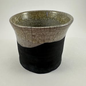 May include: A handmade ceramic cup with a unique design. The top half features a light beige, crackled glaze, while the bottom half is a solid black. The cup has a slightly flared rim and a cylindrical shape, perfect for beverages.