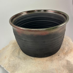 May include: A handmade ceramic pot with a dark gray exterior and a textured, ribbed design. The rim features a gradient of colors, including red, green, and gold. The pot is sitting on a beige cloth.