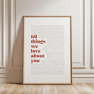 Op de afbeelding: Ingelijste print met de tekst "60 dingen die we van je houden" in rode letters. De print zit in een houten lijst en wordt tegen een witte muur getoond. De tekst is omgeven door kleinere tekst.