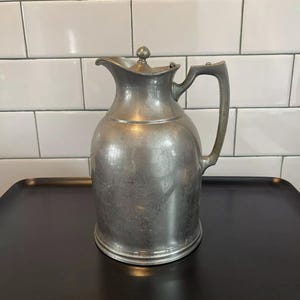Puede incluir: Una jarra vintage de color plateado con tapa y asa. La jarra tiene un cuerpo redondeado y un pico para verter. El asa es curva y est&aacute; unida al lateral. La jarra est&aacute; sobre una bandeja negra.