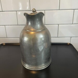 Puede incluir: Una jarra de peltre vintage con un asa decorativa y un pico curvo. La jarra tiene un cuerpo bulboso y un borde ligeramente ensanchado. Se encuentra sobre una bandeja negra contra un fondo de azulejos blancos.