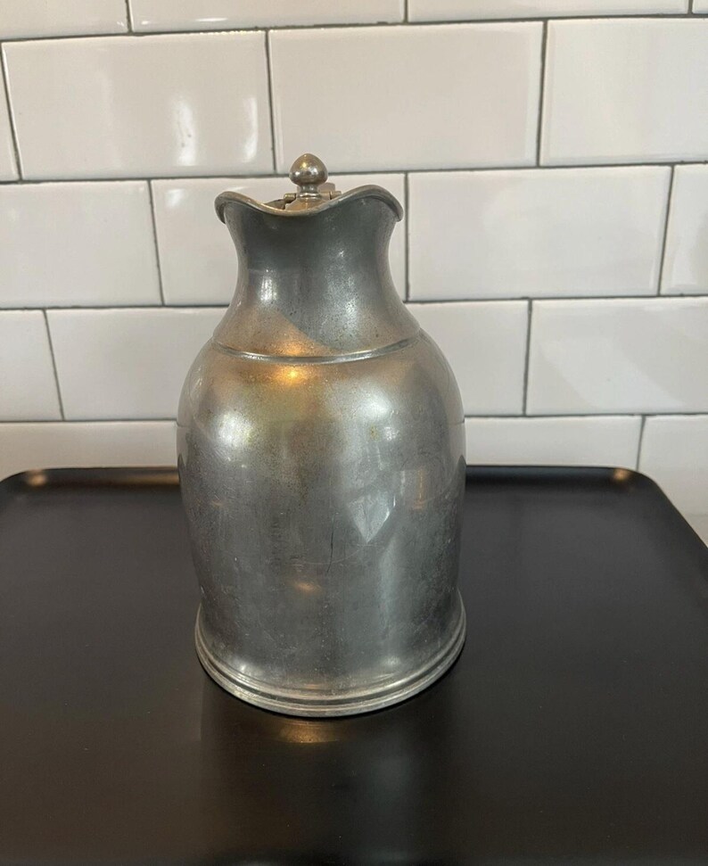 Puede incluir: Una jarra de peltre vintage con un asa decorativa y un pico curvo. La jarra tiene un cuerpo bulboso y un borde ligeramente ensanchado. Se encuentra sobre una bandeja negra contra un fondo de azulejos blancos.