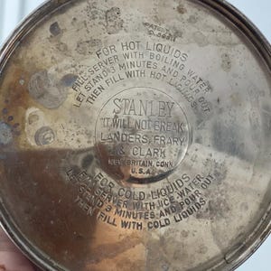 Puede incluir: Primer plano de la base de un termo Stanley vintage. La superficie met&aacute;lica plateada tiene el texto "Stanley, It Will Not Break". Tambi&eacute;n son visibles las instrucciones para l&iacute;quidos calientes y fr&iacute;os.