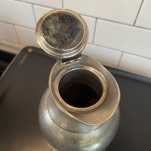 Puede incluir: Una jarra vintage de color plateado con tapa abisagrada, abierta para revelar el interior. La jarra tiene un cuerpo redondeado y un pico. La superficie muestra signos de edad y uso, lo que a&ntilde;ade encanto. La jarra est&aacute; sobre una superficie oscura.