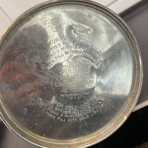 Puede incluir: Primer plano de la tapa de un termo Stanley vintage. La tapa plateada presenta texto en relieve, incluyendo "STANLEY" y "IT WILL NOT BREAK". Tambi&eacute;n se imprimen instrucciones para l&iacute;quidos calientes y fr&iacute;os.