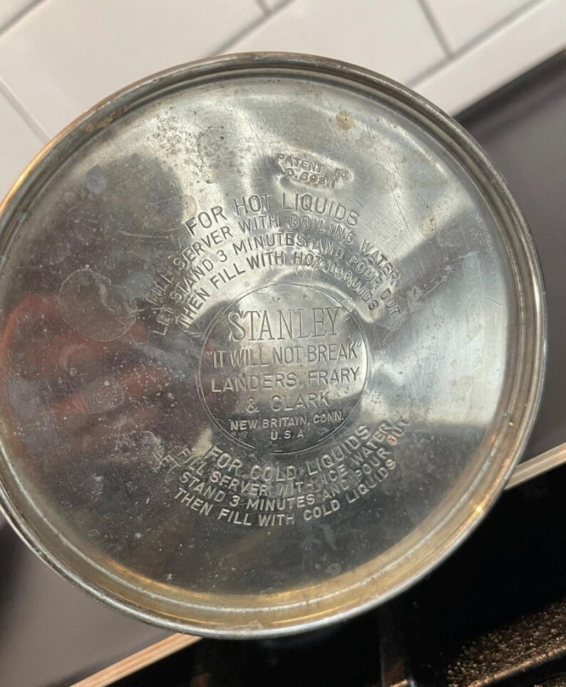 Puede incluir: Primer plano de la tapa de un termo Stanley vintage. La tapa plateada presenta texto en relieve, incluyendo "STANLEY" y "IT WILL NOT BREAK". Tambi&eacute;n se imprimen instrucciones para l&iacute;quidos calientes y fr&iacute;os.