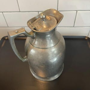Puede incluir: Una jarra de metal vintage con tapa abisagrada y asa curva. La jarra tiene un cuerpo redondeado y un pico. El metal tiene un aspecto opaco y envejecido. La jarra est&aacute; sobre una bandeja negra.