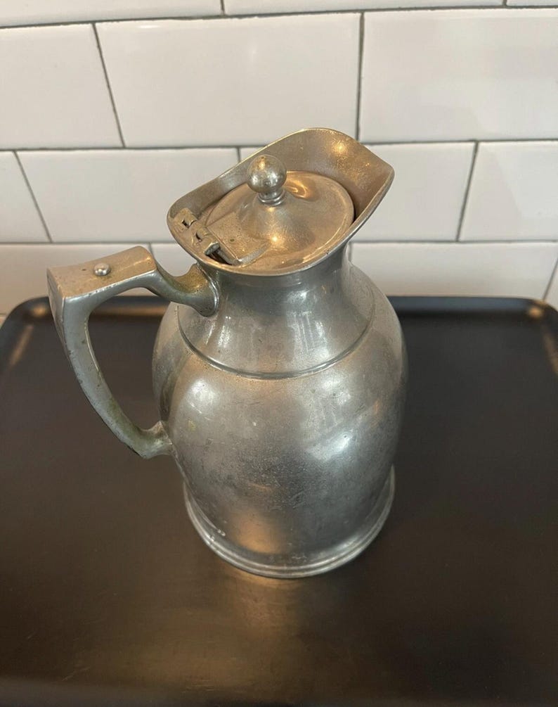 Puede incluir: Una jarra de metal vintage con tapa abisagrada y asa curva. La jarra tiene un cuerpo redondeado y un pico. El metal tiene un aspecto opaco y envejecido. La jarra est&aacute; sobre una bandeja negra.