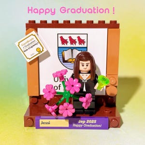 May include: A Lego graduation scene featuring a minifigure holding pink flowers and a green object. The scene includes a certificate, a university crest, and a sign that reads "Happy Graduation!" and "Sep 2025".