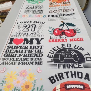 May include: A large white sheet featuring multiple colorful graphic designs including an Eiffel Tower, a turkey, cherries, a video game controller, a football with the name Nicolas, and various text phrases like I Gave Birth 21 Years Ago and Grandma.