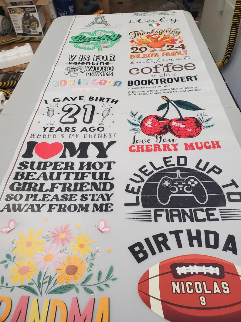 May include: A large white sheet featuring multiple colorful graphic designs including an Eiffel Tower, a turkey, cherries, a video game controller, a football with the name Nicolas, and various text phrases like I Gave Birth 21 Years Ago and Grandma.