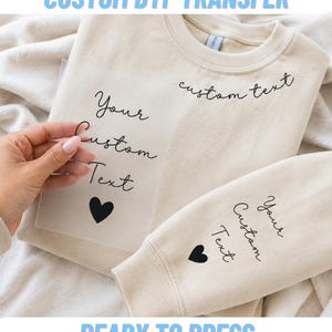 May include: A beige crewneck sweatshirt featuring black script text that reads Your Custom Text with a heart icon. A clear DTF transfer sheet with the same design is held over the fabric, showing how the custom text and heart appear when applied.