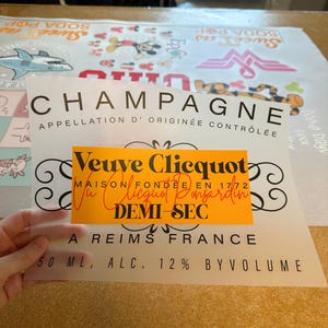 May include: A translucent rectangular sheet featuring a black printed champagne label design with the text CHAMPAGNE APPELLATION D' ORIGIN&Eacute;E CONTR&Ocirc;L&Eacute;E, a central orange rectangle with Veuve Clicquot MAISON FOND&Eacute;E EN 1772, and 50 ML, ALC. 12% BY VOLUME.
