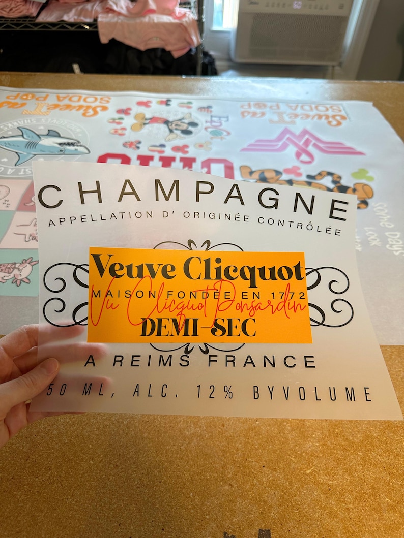 May include: A translucent rectangular sheet featuring a black printed champagne label design with the text CHAMPAGNE APPELLATION D' ORIGIN&Eacute;E CONTR&Ocirc;L&Eacute;E, a central orange rectangle with Veuve Clicquot MAISON FOND&Eacute;E EN 1772, and 50 ML, ALC. 12% BY VOLUME.