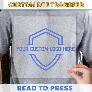 May include: A gray t-shirt with a custom DTF transfer. The transfer features a blue geometric design with the text "YOUR CUSTOM LOGO HERE." The words "CUSTOM DTF TRANSFER" and "READ TO PRESS" are also visible.