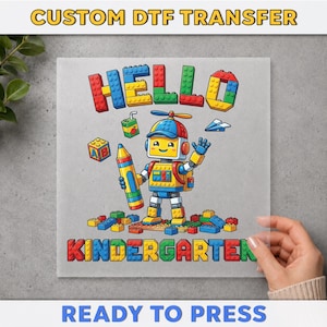 May include: A custom DTF transfer featuring a cartoon robot wearing a propeller hat, holding a large yellow crayon, and waving. The design includes the text HELLO KINDERGARTEN in colorful block-style letters, scattered building bricks, and a juice box.