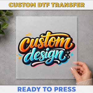 May include: A square transparent sheet featuring the text CUSTOM DTF TRANSFER at the top and READY TO PRESS at the bottom. The center displays the words custom design in a stylized, bold, graffiti-inspired font with orange and blue gradients.