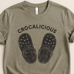 May include: Heather olive green t-shirt with the word "CROCALICIOUS" above a graphic of two black Crocs. The shirt is a Bella+Canvas brand.