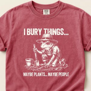 May include: Crimson red t-shirt with the text "I BURY THINGS... MAYBE PLANTS... MAYBE PEOPLE." A frog wearing overalls and a hat is depicted digging with a shovel. The shirt is a Comfort Colors brand.