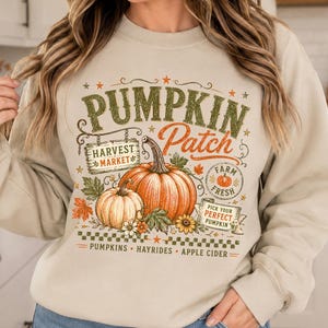May include: A beige crewneck sweatshirt featuring a graphic print with the text PUMPKIN PATCH, HARVEST MARKET, FARM FRESH, and PICK YOUR PERFECT PUMPKIN. The design includes two orange pumpkins, autumn leaves, sunflowers, and the words PUMPKINS, HAYRIDES, APPLE CIDER.
