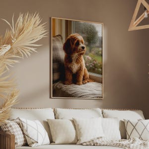 Op de afbeelding: Een ingelijste wandkunstprint met een goudbruine hond met krullend haar die op een gestructureerde deken bij een raam zit. De hond kijkt naar rechts, met een regenachtig buitentafereel zichtbaar door de ruit achter het dier.