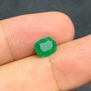 May include: A bright green emerald gemstone with a softly rounded rectangular form. The gem features a faceted surface, reflecting light. The emerald is held between fingers, highlighting its colour and clarity.