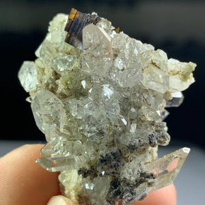 Puede incluir: Un grupo de cristales de cuarzo transparente con una inclusión mineral oscura y rectangular. Los cristales de cuarzo son translúcidos y tienen terminaciones afiladas y puntiagudas. La base es de color beige claro con depósitos minerales gris oscuro y negro.
