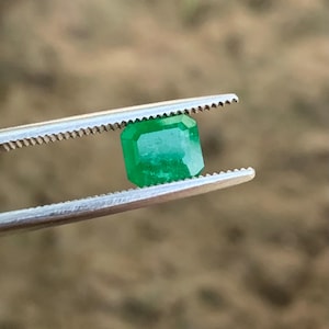 May include: A vibrant, emerald-cut green gemstone is held by a pair of silver tweezers. The gemstone has a rectangular shape with beveled edges and a bright, reflective surface. The background is blurred, suggesting an outdoor setting.