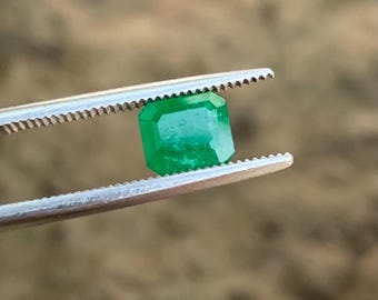 May include: A vibrant, emerald-cut green gemstone is held by a pair of silver tweezers. The gemstone has a rectangular shape with beveled edges and a bright, reflective surface. The background is blurred, suggesting an outdoor setting.