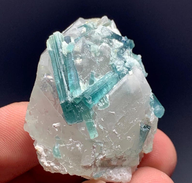 May include: A close-up of a mineral specimen featuring a cluster of teal-colored tourmaline crystals embedded in a clear quartz matrix. The crystals are elongated and have a glassy appearance, with a few small crystals scattered around the main cluster.