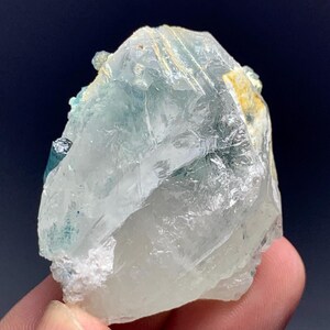 May include: A translucent, crystalline mineral specimen with pale blue and clear sections. The rough-hewn stone has a textured surface and a few small, darker blue inclusions. The mineral is held in a hand against a dark background.
