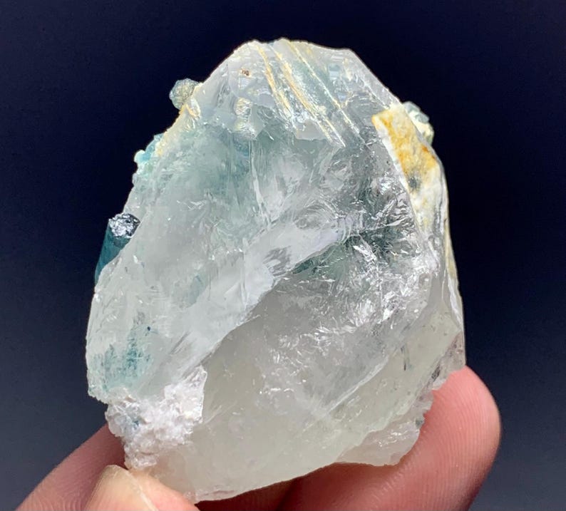 May include: A translucent, crystalline mineral specimen with pale blue and clear sections. The rough-hewn stone has a textured surface and a few small, darker blue inclusions. The mineral is held in a hand against a dark background.