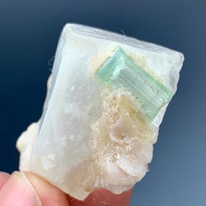 May include: A mineral specimen showing a large, translucent, light-coloured crystal with a rectangular, light green crystal embedded within. The base of the specimen has a rough, beige texture. The background is a blurred dark blue.