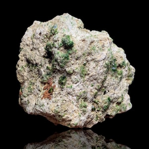 Pode incluir: Uma rocha áspera, de cor bege, com cristais verdes embutidos e algumas manchas de material castanho-avermelhado. A rocha está sobre um fundo preto, com o seu reflexo visível por baixo.