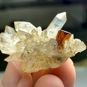 May include: A cluster of clear quartz crystals with a prominent, sharp-edged, reddish-brown crystal. The quartz crystals are translucent and clustered together, with a rough, earthy base. The overall color palette is a mix of clear, brown, and off-white.
