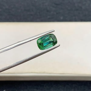 May include: A vivid green gemstone, held by silver tweezers. The gem has a rectangular cushion cut with rounded corners, displaying a rich, emerald hue with hints of blue. The polished surface reflects light, showcasing its clarity and brilliance. A close-up view of a precious stone.