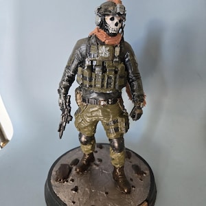 May include: A painted figurine of a soldier wearing a skull mask, tactical helmet, olive green combat gear, and a vest. The figure holds a small firearm and stands on a circular base with the word GHOST embossed on a metallic plaque at the front.